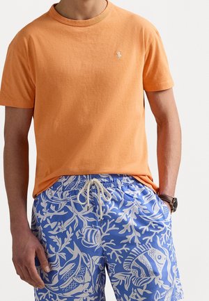 Pomarańczowa bawełniana koszulka z okrągłym dekoltem i małym haftowanym logo, zestawiona z niebieskimi kąpielówkami w wzór koralowców i ryb.