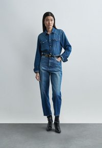 Γυναίκα στέκεται μπροστά σε λευκό απλό τοίχο, φορώντας εφαρμοστό τζιν πουκάμισο, ψηλόμεσα τζιν, μαύρη ζώνη και μαύρα μποτάκια μέχρι τον αστράγαλο.