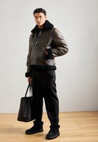 Giacca bomber in pelle marrone con colletto e polsini in shearling nero, abbinata a pantaloni neri e una borsa tote nera. Finitura testurizzata.