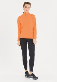 Oranssi pitkähiha pulloveri, jossa on puolivetoketju, yhdistetty mustiin legginsseihin ja mustiin urheilukenkiin, joissa on harmaa ja vaaleanpunainen pohja.