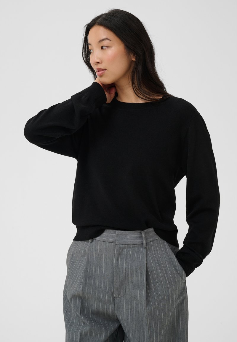 Femme aux longs cheveux foncés portant un pull noir et un pantalon gris à fines rayures, regardant sur le côté avec une main reposant près de son cou.