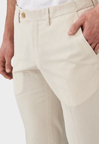 Pantalones de color beige claro, texturizados y con un patrón acanalado. Cuentan con un cierre de botón y bolsillos laterales, mostrando un ajuste a medida.