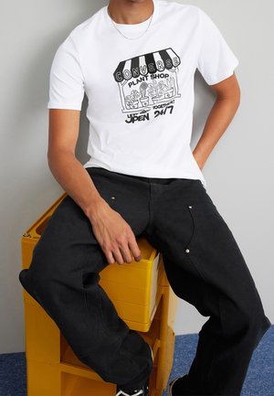 Homme portant un t-shirt blanc avec une illustration d'une boutique de plantes noire et du texte, pantalon noir, assis sur des caisses jaunes empilées contre un mur gris.