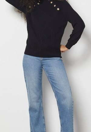 Femme portant un pull en maille noir avec des boutons dorés et un jean bleu clair à jambes droites, posant avec une main sur la hanche.