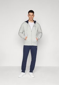 Siva zip-up dukserica s mornarskim akcentima, prednjim džepovima i bijelom majicom ispod. Uparena s mornarskim trenirkama i bijelim tenisicama.