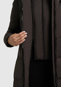 Dunkelbraune Puffjacke mit einer gesteppten Textur, ausgestattet mit mehreren Reißverschlüssen und einer taillierten Innenschicht. Eine Hand wird gezeigt, die den Reißverschluss hält.