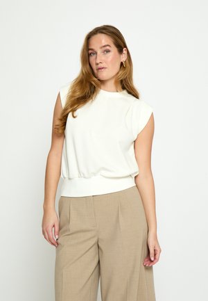 Naine pikkade helepruunide juustega, kes kannab valget varrukateta toppi ja beeže laia säärega pükse, seisab ühetaolise heleda tausta ees.