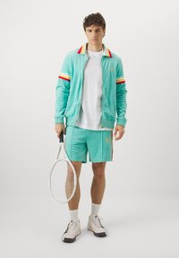 Türkiissinine veluurtreeningkostüüm punaste ja kollaste triipudega, valge t-särk, sobivad lühikesed püksid, hoiab tennisereketit, jalas heledad tossud.