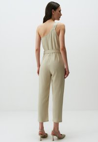 Tutina beige con spalla unica, vita con cintura, gambe affusolate e texture liscia. Abbinata a scarpe con tacco alto, con cinturini e texture. 