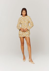 Blazer giallo a scacchi con gonna abbinata, caratterizzato da un design strutturato, maniche lunghe e bottoni. Indossato con scarpe beige con tacco alto.
