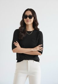 Czarny sweter z krótkim rękawem z teksturowanymi motywami serc, w połączeniu z kremowymi spodniami i ciemnymi okularami przeciwsłonecznymi. Prosty złoty naszyjnik jako dodatek.