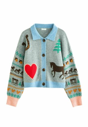Jeune fille aux longs cheveux portant un cardigan gris avec des motifs colorés, un jean bleu et des chaussures marron, se tient contre un mur blanc en souriant.