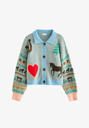 Jeune fille aux longs cheveux portant un cardigan gris avec des motifs colorés, un jean bleu et des chaussures marron, se tient contre un mur blanc en souriant.
