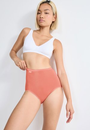 Giovane donna con capelli corti e biondi, indossa un reggiseno bianco e mutandine corallo a vita alta, posa contro uno sfondo chiaro e uniforme.