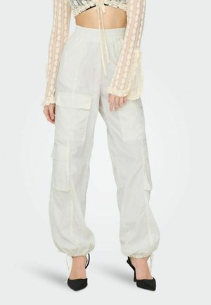 White cargo pants with multiple pockets, drawstring hem, and elastic waistband, paired with a sheer long-sleeve top and black heels.