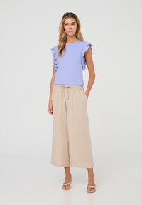 Top corto lilla a maniche corte con dettagli arricciati in pizzo, abbinato a pantaloni beige a gamba larga e sandali neutri, creando un outfit leggero e confortevole.