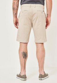 Beige Baumwollshorts mit einem entspannten Schnitt, Seiten- und Gesäßtaschen sowie einer glatten Textur. Getragen mit grauen Leinenschuhen. Palmbaum-Tattoo sichtbar.