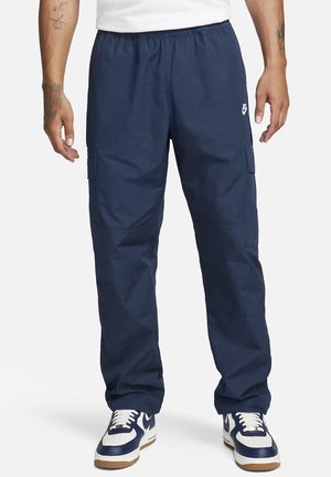 Homme portant un pantalon cargo bleu marine avec une taille élastique et des baskets blanches et bleu marine, debout devant un fond uni.