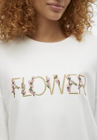 Vit sweatshirt med "FLOWER" i guldtrådsbroderi, accentuerad med rosa blommönster, mot en slät tygtextur.