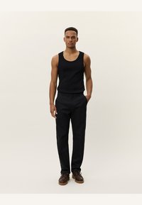 Zwarte tanktop en op maat gemaakte zwarte broek met een rechte snit. Bruine leren schoenen met veters en textuur op de zolen.