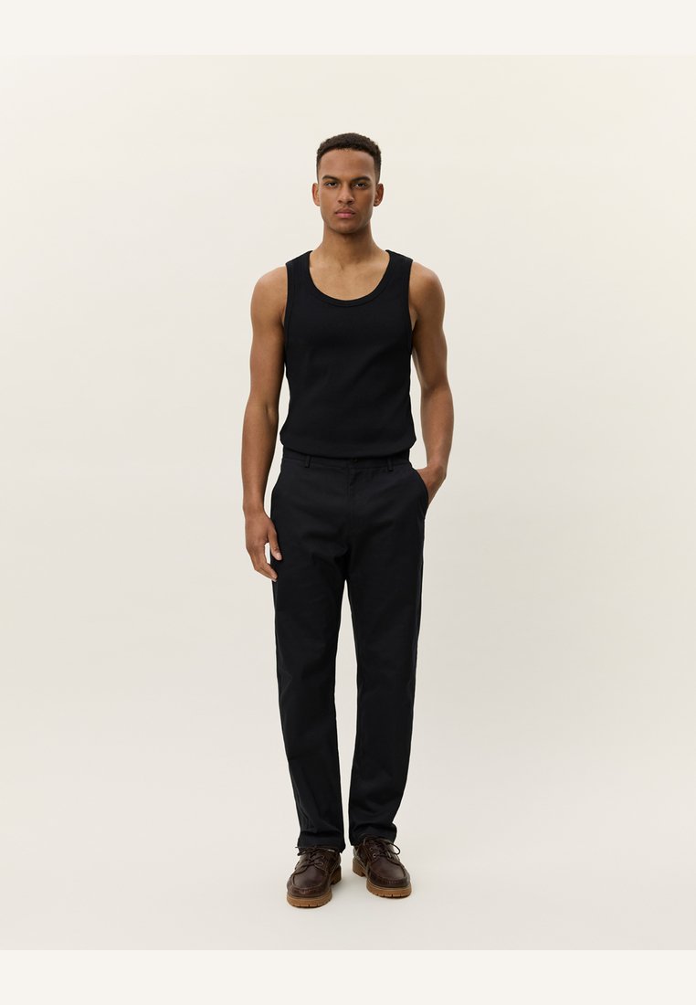 Zwarte tanktop en op maat gemaakte zwarte broek met een rechte snit. Bruine leren schoenen met veters en textuur op de zolen.