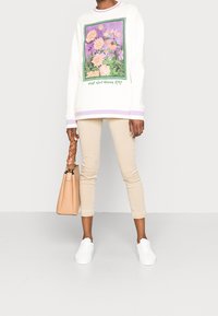 Sudadera blanca con estampado floral y detalles en púrpura; pantalones cortos beige; zapatillas blancas; bolsa marrón con asa trenzada. Atuendo simple y casual.