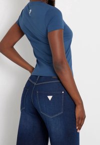 Blå tætsiddende t-shirt med korte ærmer vist sammen med højtaljede mørke denimjeans med trekantet mærkedetalje på baglommen.