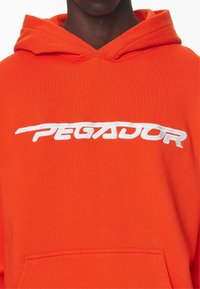 Oranger Kapuzenpullover mit einer Fronttasche, verziert mit weier gestickter Schrift "PEGADOR" auf der Brust. Weiche Stoffstruktur.