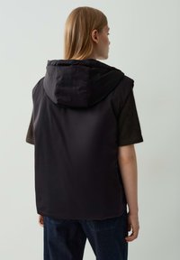 Frau mit glattem, blonden Haar, die eine schwarze ärmellose Kapuzenweste über einem dunklen Kurzarmoberteil und dunkelblauen Jeans trägt, die sich abwendet.