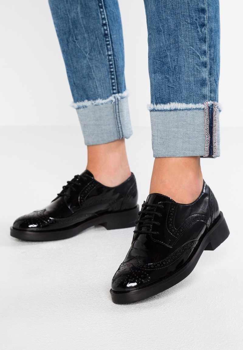 Chaussures derby en cuir verni noir avec des détails brogue décoratifs, un design à lacets et un talon épais, associées à un jean bleu retroussé.