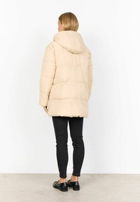 Chaqueta acolchada beige con capucha, que presenta un acolchado de rombos, un corte relajado y un dobladillo ajustable, combinada con jeans negros ajustados y mocasines.