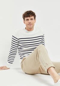 Maglione bianco con righe navy, scollatura rotonda e tre bottoni sulla spalla sinistra. Pantaloni beige, seduto su uno sfondo semplice.