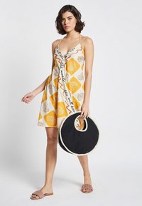 Vestido amarillo y crema con patrones florales y ondulados, escote en V y tirantes finos, combinado con un bolso tote negro circular.