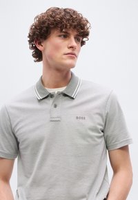 Jeune homme aux cheveux bouclés portant un polo gris clair avec un col rayé blanc et le logo "BOSS" sur la poitrine, regardant sur le côté.