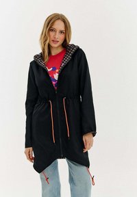 Melna zipa jaka ar pārbaudītu kapuci, oranžiem stringiem un asimetrisku malas griezumu. Saskaņota ar sarkanu grafisko T-kreklu un gaišām džinsām.