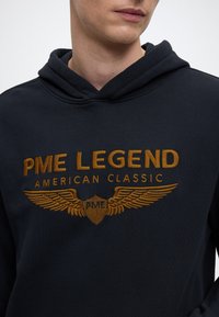 Črna kapuca z zlatimi vezenimi napisi "PME LEGEND AMERICAN CLASSIC" in grafikonom kril. Mehka tekstura, priložnostna zasnova.