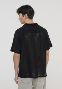 Maglietta nera a maglia con colletto corto, caratterizzata da un motivo a traforo e maniche corte, abbinata a pantaloni grigio chiaro.