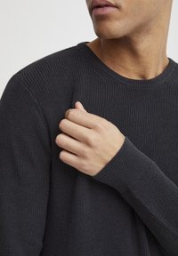 Pull en tricot gris foncé avec une texture côtelée, col rond et manches longues, comportant un détail de poignet ajusté.