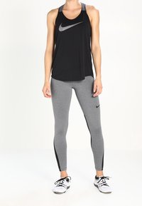Czarny bezrękawnik z dużym szarym logo Nike, zestawiony z szarymi legginsami z czarnymi paskami po bokach i białymi butami sportowymi.