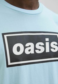 Šviesiai mėlynas medvilninis marškinėlis su juodu stačiakampiu grafiku, kuriame baltomis didelėmis raidėmis užrašyta "oasis". Klasikinio apvalaus kaklo dizainas.