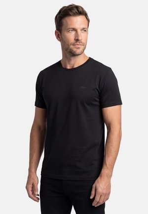 Mann mit kurzem braunem Haar und Bart, der ein schlichtes schwarzes T-Shirt mit kurzen Ärmeln trägt und seitlich vor einem weißen Hintergrund blickt.