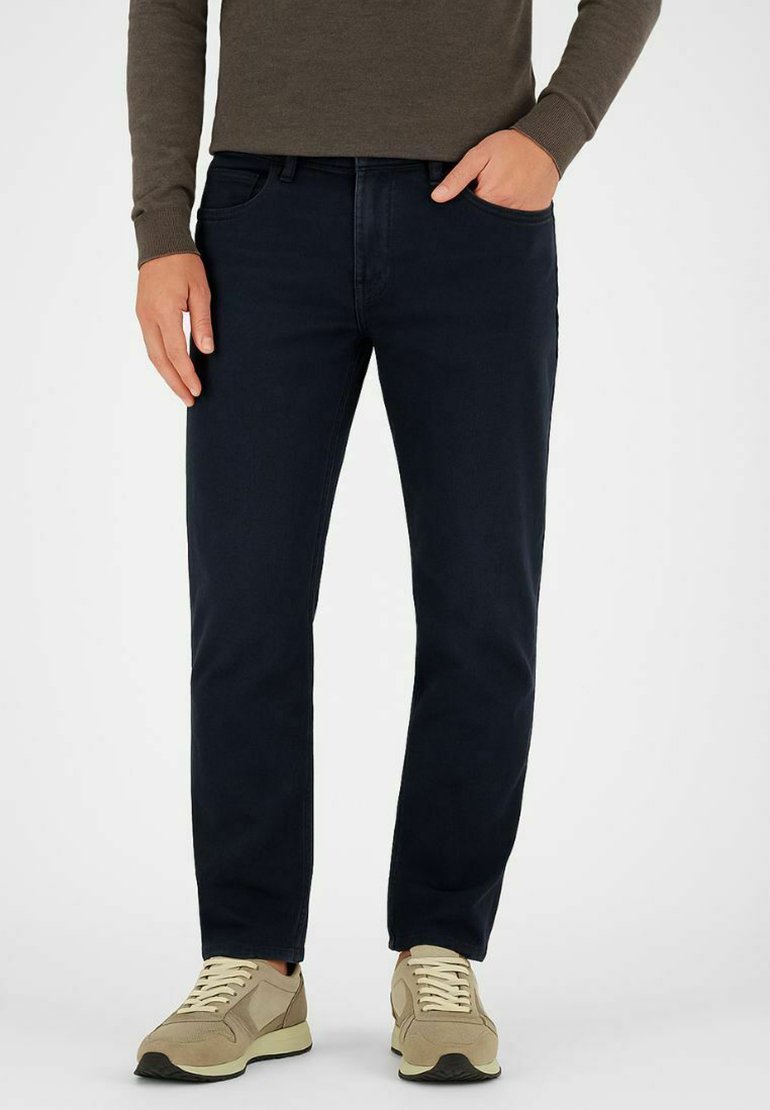 Dunkelblaue Jeans mit slim fit, aus Baumwoll-Mischmaterial und im fünf-Taschen-Design. Kombiniert mit beigefarbenen Sportschuhen und einem braunen Langarmshirt.