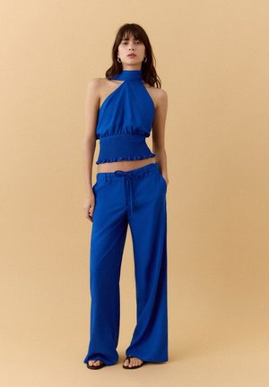 Mujer con top corto halter azul cobalto y pantalones anchos a juego, de pie contra un fondo beige con las manos en los bolsillos.