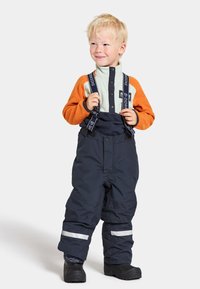 Marina vattentäta snöoverall med reflexränder, justerbara hängslen och en mjuk grön och orange fleeceöverdel. Barnet har på sig svarta stövlar.