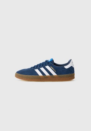 Sininen mokkanahkainen Adidas-tennari, jossa on valkoiset raidat, kumipohja ja "Montreal" kultaisella tekstillä sivulla, kuvattuna vasemmalta puolelta valkoisella taustalla.