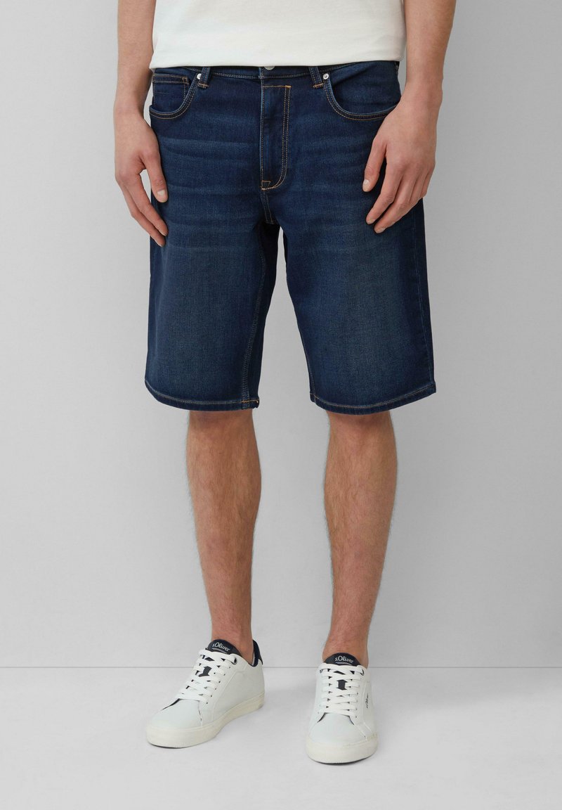 Shorts en denim bleu foncé avec une coupe droite, un design à cinq poches et des coutures orange contrastantes. Portés avec des baskets blanches.