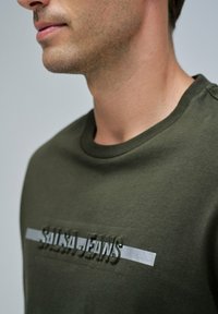 Primer plano de un hombre que lleva una camisa de color verde oliva con texto reflectante en relieve en el pecho.