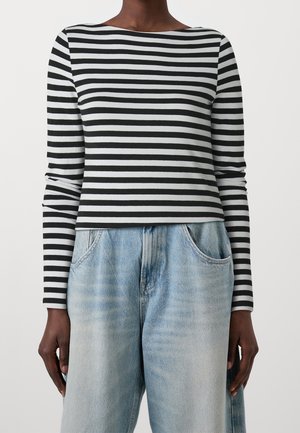 Personne à la peau foncée portant un t-shirt à manches longues rayé horizontal noir et blanc et un jean taille haute bleu clair, les mains détendues le long du corps.