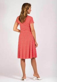 Rotes Kleid mit weißen Punkten, kurzen Ärmeln, einem taillierten Oberteil und einem ausgestellt Rock. Glattes Material, knielang, getragen mit weißen Blockabsatzschuhen.