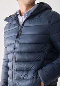 Námořnická puffer bunda s kapucí. Obsahuje horizontálně prošívané sekce, plný zip a boční kapsy. Vyrobena z lesklého, lehkého materiálu.
